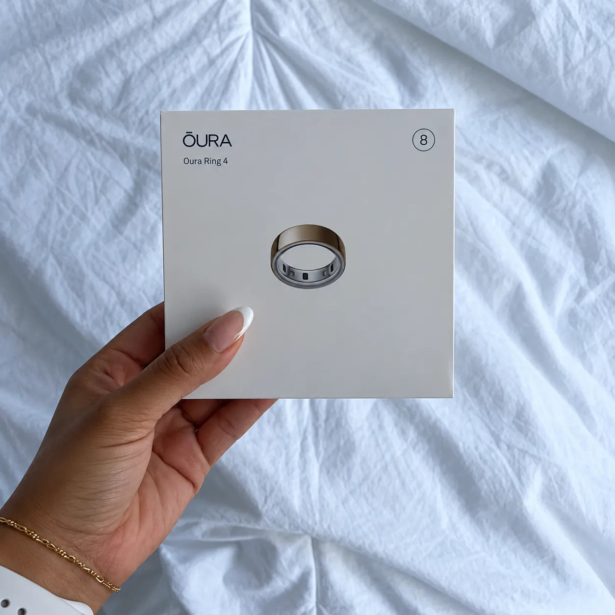 A hand holding an Oura Ring 4 box over white bed linen