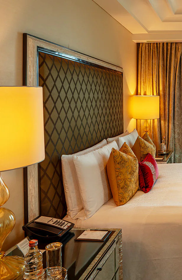 A hotel bed with gold cushions beside a lit bedside lamp